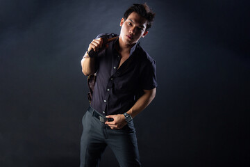Fototapeta premium An attractive Asian man dressed in all-black formal attire poses confidently in a dramatic studio setting, holding a stylish brown duffle bag with a strong and dynamic stance against a dark background