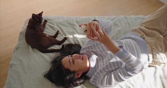 young Asian woman lies on a tidy bed using a smartphone as a brown cat lounges nearby. top view