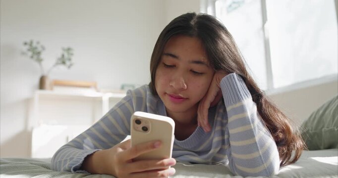 Asian happy young woman on bed using smartphone in soft morning window light at home