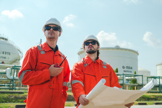 Oil and gas refinery in petroleum industry with inspection engineer, industrial worker in orange coverall and hard hat at oil tank site, holding blueprint with radio