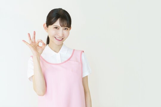 白背景のピンクのエプロンをつけた指で丸のポーズをする若い日本人女性介護士のポートレート