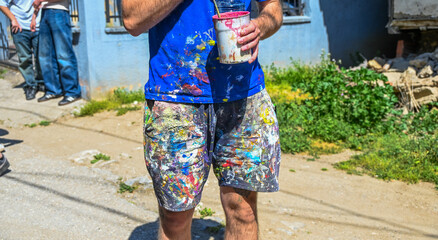 A man prepares paint for a mural. An artist in shorts and a T-shirt smeared with paint. Outdoor mural painting. © Vanja Stokic
