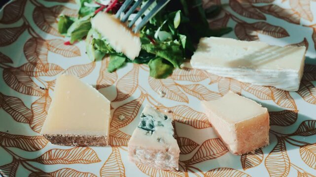 Person cutting pieces of gourmet cheese on patterned plate with salad greens.