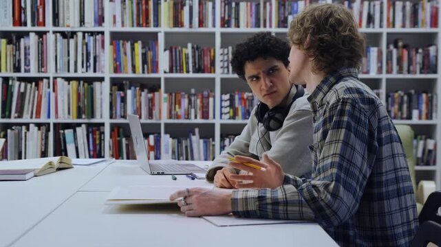 A friendly study session unfolds as two students work side by side on homework. The atmosphere feels relaxed yet productive, with a girl in the background selecting a book among wooden shelves.