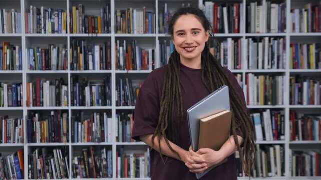 Positive young woman with dreadlocks smiling happily while holding a laptop and a book in a library