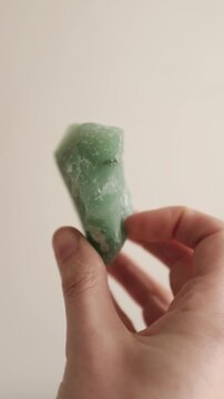 Close-up of a person's hand holding and turning a raw, unpolished green aventurine quartz crystal gemstone against a neutral studio background, vertical orientation.