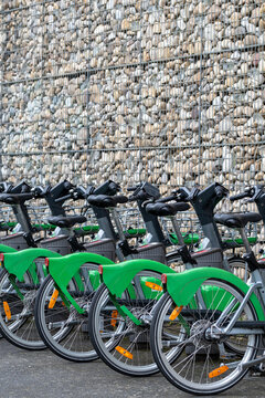 Bikeshare bicycles line a Lyon station in a rental row by the sidewalk, where wall texture, mobility and transport feel ordered and still.