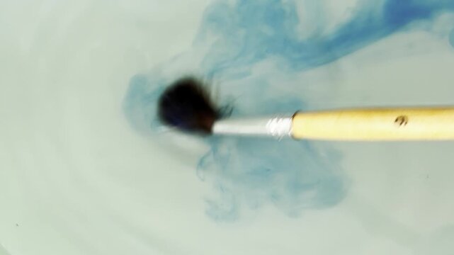 Artist rinsing a brush with blue watercolor paint in a jar of water creating a fluid abstract swirl. Creative process of dissolving ink for an artistic effect