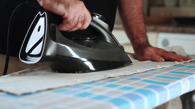 Man carefully ironing wrinkled fabric with a black iron on a patterned board