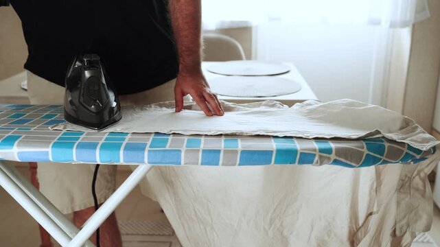 Man irons clothes with black iron on blue board at home during day