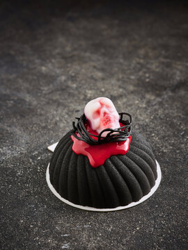 This dessert showcases a black, textured pastry topped with a skull and red sauce, designed for Halloween festivities