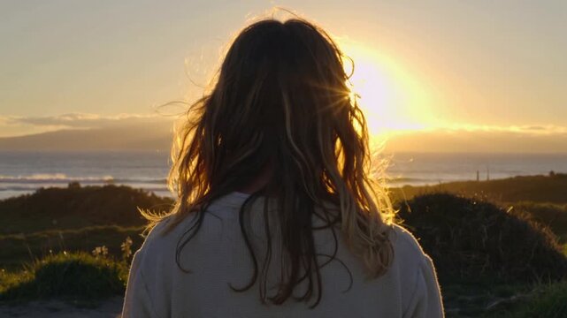 Watching woman standing, watching sunset at ocean on dunes, sun brightening hair and light shirt