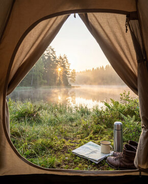 A mug and thermos sit at the tent entrance facing a quiet lake, showing a screen-free camping morning and anticipation of travel.