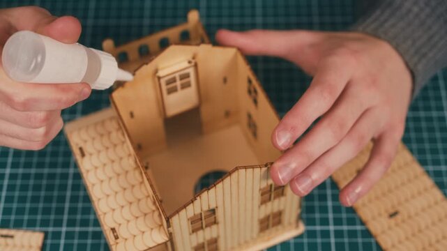 Assembling wooden house model with hands positioning roof section, applying glue for DIY miniature home construction. New housing concept, creative architecture hobby, handmade property prototype