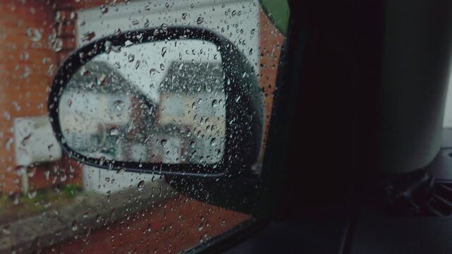 Raindrops covering parked car side mirror near quiet suburban street during heavy rain. Waiting inside stationary vehicle with wet window creating moody atmospheric weather concept. Stormy