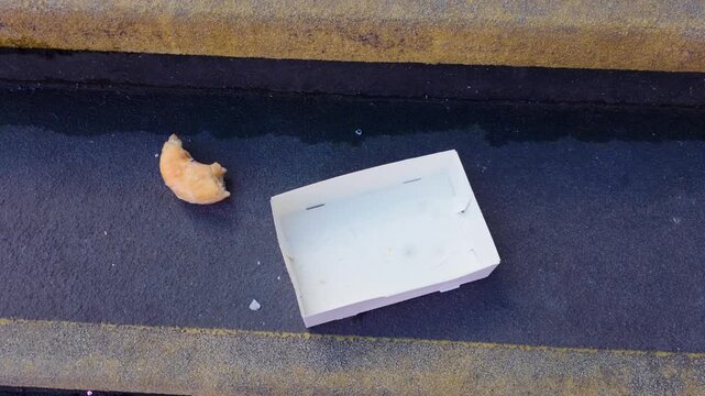 Half eaten donut beside empty takeaway box on concrete steps in urban setting. Discarded food waste suggesting poverty homelessness street survival and social inequality concept. City neglect and