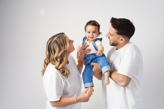 A cheerful family with a happy, baby. The parents are smiling and looking lovingly at their daughter. Everyone is wearing casual white tops and jeans.
