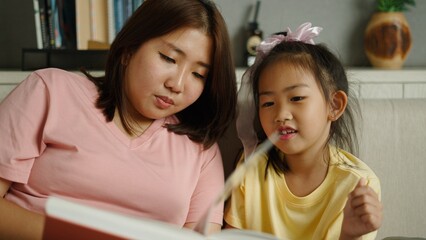 Obraz premium Asian mother and daughter reading book together at home, learning and bonding