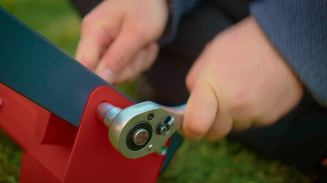 Hands operating ratchet wrench securing structural connection on painted metal frame. Tool-driven assembly process with visible bolt and red mounting plate. Home improvement and equipment setup theme