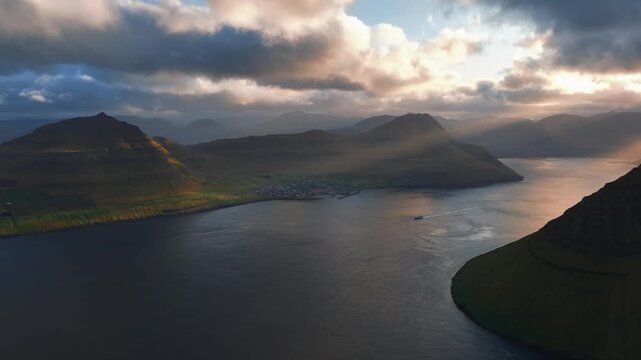 Mountain fjord landscape in the Faroe Islands with dramatic stratocumulus clouds, low golden sunlight, ocean inlet and green grassy slopes during late summer evening, slow orbit approach drone shot