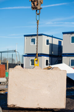 Construction site shows a crane lifting a heavy concrete block near worker cabins under a clear blue sky