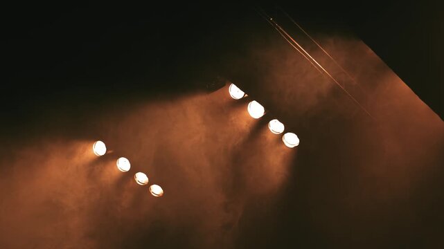 Dramatic low angle shot of theater or concert stage lights. Warm amber spotlights shining through dense haze and artificial smoke on a dark background. Perfect for artistic backgrounds and theatrical 