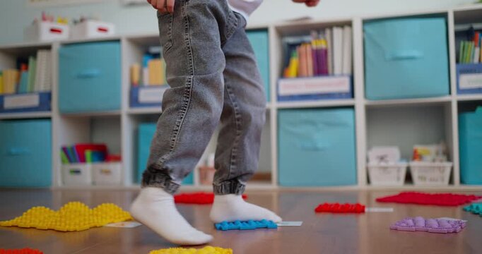 Child in white socks walking across colorful textured sensory mats on a classroom floor, practicing balance and tactile play near storage cubbies