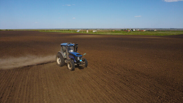 A high-angle drone shot of a blue tractor pulling farm equipment across a vast, dark field. A thick cloud of dust rises from the dry soil as the machinery prepares the land for planting under a clear 