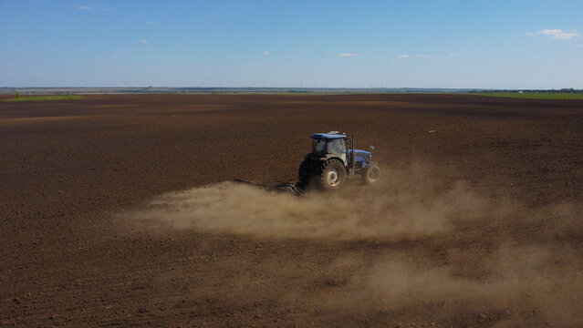 A high-angle drone shot of a blue tractor pulling farm equipment across a vast, dark field. A thick cloud of dust rises from the dry soil as the machinery prepares the land for planting under a clear 