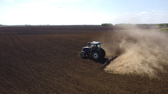 A high-angle drone shot of a blue tractor pulling farm equipment across a vast, dark field. A thick cloud of dust rises from the dry soil as the machinery prepares the land for planting under a clear 