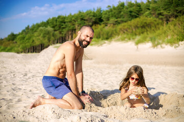 Fototapeta premium Happy bearded father playing in the sand with his little daughter wearing sunglasses on a sunny beach, concept of family summer vacation and paternal bond
