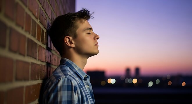young man with eyes closed leans against a brick wall, finding peace as the sun sets and city lights twinkle in the distance.