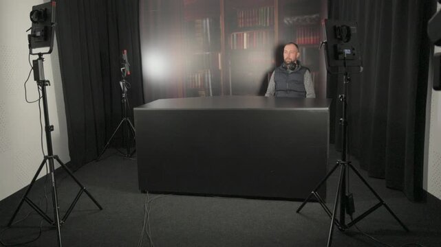 A man sitting at a desk in a studio or home office, scrolling through a smartphone app or texting against a library bookshelf background.