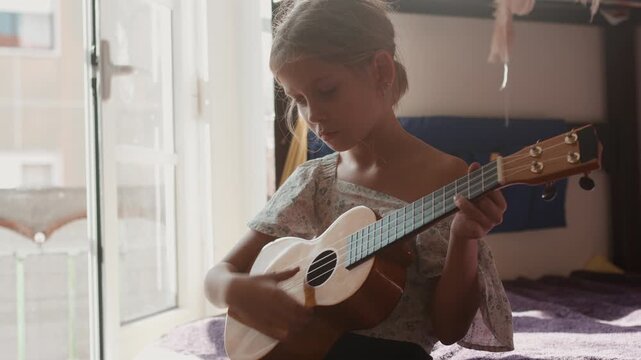 Young girl playing ukulele near window at home. Child holding string instrument and focusing while practicing music at home