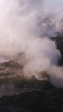 Aerial view of Victoria Falls at dusk in Zimbabwe shows rapids, falling water, billowing spray, wooded banks, small islands, and village hints in warm golden pink light.