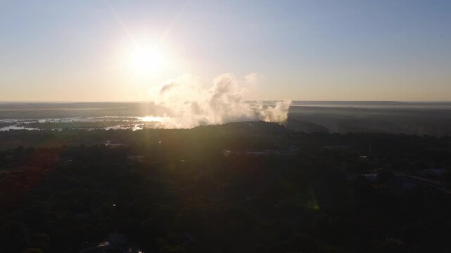 Wide aerial view shows Victoria Falls in Zimbabwe as mist and spray drift upward. The Zambezi River, riverside village, gorge, and faint bridge silhouette reflect golden light.
