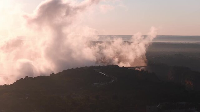 Aerial view shows Victoria Falls on the Zambezi River in Zimbabwe as mist surges upward and drifts, with Victoria Falls Bridge, a winding road, and village silhouettes.