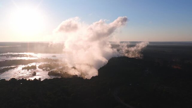 Aerial dusk view shows Victoria Falls, Zambezi River, and gorge in Zimbabwe. Golden hour backlights spray as mist billows and drifts over islands and village rims.