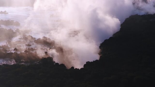Aerial sunset view of Victoria Falls in Zimbabwe shows the Zambezi River, rocky channels, gorge rim, islands, and braided rapids as a white spray plume billows.