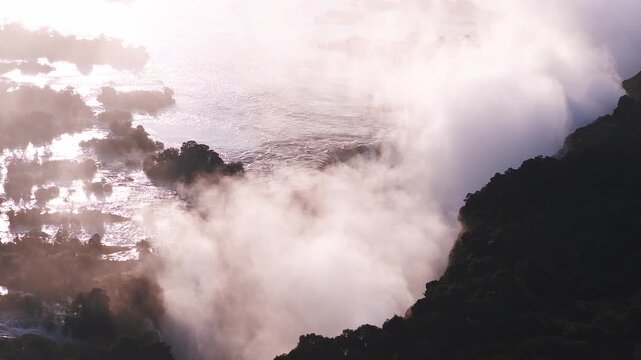 Aerial at Victoria Falls on the Zambezi River, Zimbabwe and Zambia, as water races to the brink and a curtain falls, with mist rising and drifting in golden light.