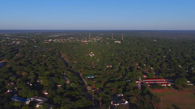 Aerial view near Victoria Falls, Zimbabwe shows treed townscape, radio towers, red roof long buildings, sports field, dirt roads, and slow forward pan in warm sunset light.
