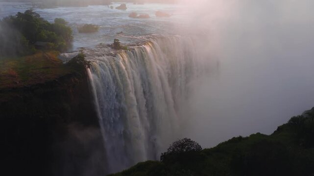 Aerial view shows Victoria Falls on the Zimbabwe side at sunset. Water plunges into a misty gorge, spray rises, islands and rapids appear, trees and a riverside clearing show scale.