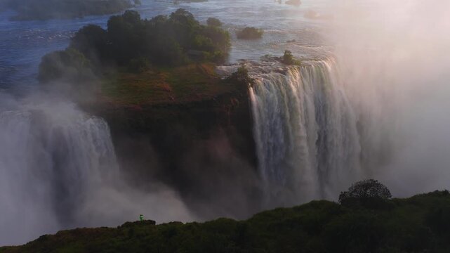 Aerial view shows Victoria Falls at sunset on the Zimbabwe Zambia border. A lone figure stands on the rim as cascades plunge, mist rises, and warm light glows.