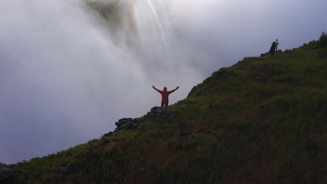Aerial sunset view shows Victoria Falls in Zimbabwe as water plunges into mist. A person in orange raises arms while two in raincoats take photos on the rim.