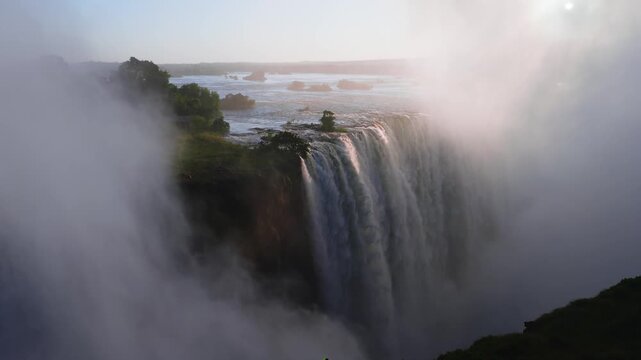 Aerial sunset shows Victoria Falls in Zimbabwe, Zambezi River islets, deep mist filled gorge, torrents cascade, currents swirl, mist drifts, a tiny person stands on rim.