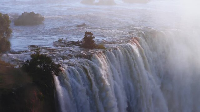 Aerial sunset view shows Victoria Falls on the Zambezi in Zimbabwe. A slow drone sweep follows the flow, silky curtains cascade, mist towers, rapids churn.