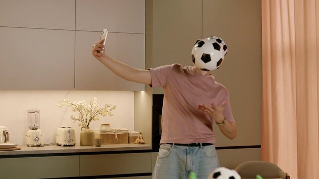 Young male soccer enthusiast in casual attire takes a selfie while juggling soccer balls in a modern kitchen with stylish decor and appliances visible in the background