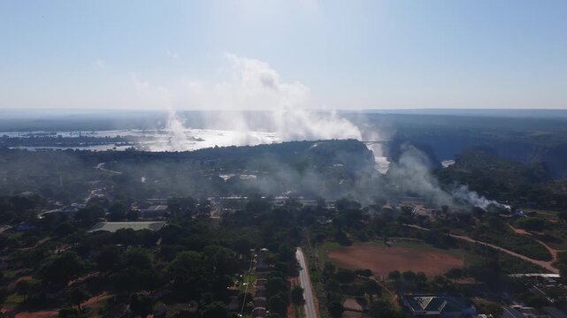 Aerial view shows Victoria Falls on the Zambezi River at sunset, the bridge, rising spray, a Zimbabwean village, a straight road, and smoke in backlit panoramic light.