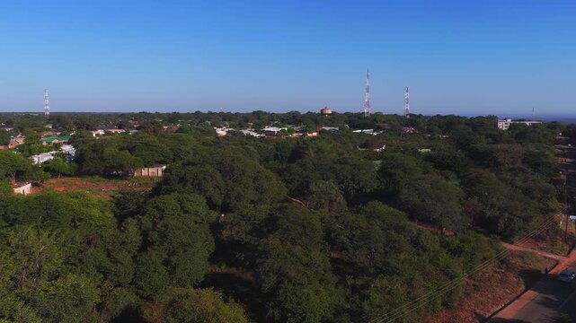 Aerial view of a tree canopied village near Victoria Falls in Zimbabwe. A white vehicle moves on a red dirt road by radio masts and a round water tower in late afternoon light.