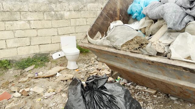A giant metal bin overflows with bags and construction debris outdoors. The unpleasant, chaotic view features a discarded white toilet and loose garbage scattered across the dusty ground.
